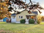 A single-story suburban house with beige siding, a dark roof, and white trim under a sunset-lit sky, featuring a wooden deck and a lawn with a large bush near the entrance.