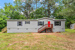 A single-story gray mobile home with a red door and wooden stairs, set against a backdrop of trees and a partially grassy yard.