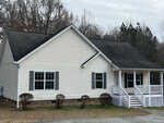A single-story white siding house with a gabled roof, featuring a small front porch with white railings, and two visible windows. the house is surrounded by bare shrubs and a partly cloudy sky overhead.