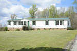 A single-story white modular home with a green roof and partial brick foundation, positioned on a grassy plot with a backdrop of trees under a partly cloudy sky.