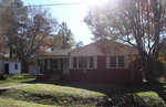 A single-story red brick house with white trim and a small porch under a clear blue sky, surrounded by green grass and trees with autumn foliage.
