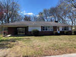A single-story brick house with a large front yard and bare trees in the background on a sunny day.