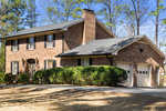 A two-story brick suburban house with a single-car garage and a landscaped front yard with bushes and bare trees under a clear sky.