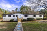 A white manufactured home with a raised foundation and a set of wooden stairs leading to the front entrance, set on a property with grass and trees under a clear sky.
