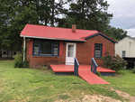 A small red brick house with a red sloped metal roof, white door, and red entrance steps, set against a backdrop of trees and a grassy lawn.