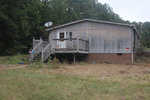 A weathered house with a wooden deck and brick foundation sits in a grassy area, with a satellite dish attached and various objects scattered on the ground.