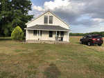 A white two-story house with a front porch and a red pickup truck parked on the right side, set against a backdrop of cloudy sky and surrounded by a green lawn.
