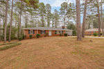 Single-story residential home with a brick facade and shingled roof, set back from the road amidst a stand of pine trees with a brown lawn in the foreground.