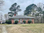 A single-story brick house with a sloped roof, surrounded by a grassy yard with tall pine trees, under a cloudy sky. a driveway leads up to the side of the house where a vehicle is parked.