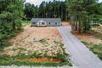 A single-story house with a gray roof and beige siding, featuring an extended driveway, is situated in a clearing surrounded by tall pine trees. the terrain appears dry with patches of grass, and a lone figure stands in the open area in front of the house.