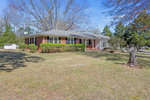 A single-story brick house with white trim, surrounded by a grassy lawn with a few trees and bushes, under a clear blue sky.