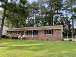 A single-story brick house with a white porch and an american flag displayed in the front yard, surrounded by tall pine trees and a well-manicured lawn.