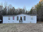 A single-story white mobile home with a black roof, featuring a central door flanked by two windows, one with a bright pink curtain, set against a backdrop of trees with a vehicle parked to the left.