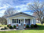 A small, single-story house with white siding and a front porch on a sunny day, with a neatly manicured lawn and bare trees in the background.