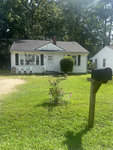 A small, single-story house with a white exterior and a black roof, featuring a front yard with a lawn and a mailbox in the foreground. there is a plastic white table and chairs on the right side of the house, and it’s a sunny day with clear skies.