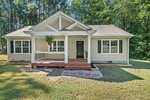 A single-story house with white siding and a gabled roof, featuring a front porch with steps and surrounded by a grassy lawn with trees in the background.