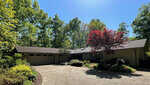 Suburban single-story house at 2108 Coleman Place, Henderson NC, with a double garage and a driveway, surrounded by green trees with one red tree near the entrance, under a clear blue sky