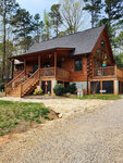 A two-story log cabin at 52 Buoy Drive, Henderson NC, with an extended porch and a staircase leading to the entrance, surrounded by trees under a clear sky.