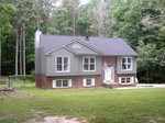 A two-story house with gray siding and red brick foundation situated in a clearing surrounded by trees at 57 Lakewood Court, Henderson NC. There is an American flag displayed on a pole attached to the