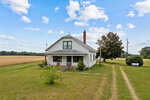 A small, white countryside house with a gabled roof and a chimney, set against a background of a clear sky and a vast, golden wheat field at 1558 Cheeks Quarter Road, Henderson