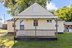 A small, two-story, beige house with a front porch and a 'for sale' sign.