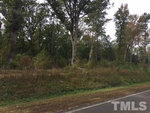 A wooded area with overgrown grass and shrubs beside a paved road in Henderson NC, with the watermark "tmls" visible.