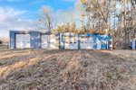A blue metal storage building with four garage doors, situated on a grassy field with trees in the background and a clear sky above, located at 1150 Gun Club Road, Henderson NC.