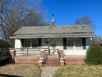 A small, white single-story house on 229 Lowry Street, Henderson NC, with a shingled roof, brick foundation, and a front porch with steps. There are bare trees and a