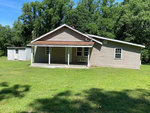 A modest single-story house with beige siding and a brown roof, featuring a small front porch, is situated on a lawn with trees in the background at 122 Bill Spain Road, Henderson NC. A
