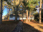 A single-story white house with a shingled roof and brick chimney, set amidst tall pine trees at 120 Cedarwood Drive, Henderson NC, with a walkway leading to the front door.