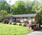 Single-story brick house with a gabled roof, front porch, and landscaped garden surrounded by lush green trees at 1257 David Avenue, Henderson NC.