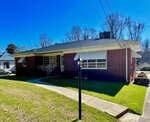Red brick single-story suburban home at 2315 Oxford Road, Henderson NC, with a sloping roof, featuring a covered front porch and a prominent chimney, under a clear blue sky. A street