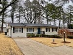 A single-story white house at 1264 Oakridge Avenue, Henderson NC, with a black roof and a blue front door, featuring a concrete driveway and surrounded by tall pine trees.