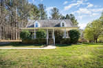A two-story suburban house at 119 Community House Road, Henderson NC, with a gabled roof, white siding, and a front porch, is beautifully set against a backdrop of tall trees and a