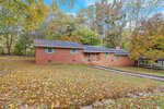 A single-story brick house at 139 Ellington Road, Henderson NC, with a black shingled roof, featuring a small porch area and surrounded by a lawn covered in fallen autumn leaves.