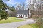 A quaint single-story house with a large chimney and white siding sits surrounded by trees on 1805 St Andrews Church Road, Henderson NC, featuring a clear front yard and a curved driveway.