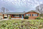A single-story brick house with a carport, located at 902 Bridgers Street Henderson NC, featuring a blue front door and surrounded by a yard with overgrown vegetation.