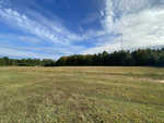 A sprawling green field at 245 Kemps Lane, Henderson NC, with patches of brown grass under a partly cloudy sky, flanked by a dense treeline and a tall tower in the distance.