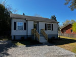 Small white single-story house at 2223 Oxford Road, Henderson NC, with gray shingles and wooden front steps, featuring two windows visible at the front and a gravel area in the foreground, with
