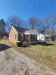 A single-story brick house with a white door and shutters, featuring a small front porch, is shown on a sunny day with a clear blue sky at 903 Dabney Drive, Henderson NC