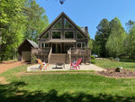 A two-story house at 147 Anchor Drive, Henderson NC, featuring a large central window and a deck with four chairs, surrounded by lush green trees on a bright sunny day.