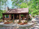 A log cabin with an American flag in a woodland setting, featuring a stone pathway, flower beds, and a patio area with outdoor furniture at 49 Pebble Hill Lane, Henderson NC.