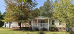 A single-story beige house with a white front porch and an American flag, located at 203 Ellington Path, Henderson NC, is surrounded by trees with early autumn foliage.