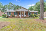 A single-story brick house at 1214 Anne Street, Henderson NC, with a centered front door and symmetrically positioned windows under a gabled roof, surrounded by a lawn with scattered trees.