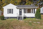 A single-story white house with black shutters and a black front door, located at 1801 Willow Lane, Henderson NC, featuring a small front porch and a pathway leading up to it, surrounded