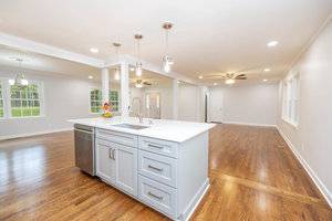 A spacious, empty kitchen featuring a large island with a sink, hardwood floors, pendant lights, and a view into adjoining rooms with multiple windows.