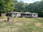 A single-story residential home with a beige exterior and brown roof, featuring a front porch and a separate brick structure in the foreground, is situated in a grassy yard with trees in the background, located