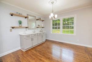 A bright, empty room featuring a white vanity with a mirror, flanked by two wooden shelves, and a large window providing natural light.