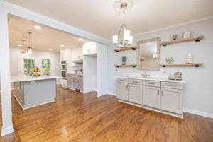 A spacious, well-lit kitchen with hardwood floors, featuring an island, white countertops, and wooden shelves against a white wall.
