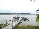 A metal walkway at 110 Somerset Lane, Henderson NC, leads to a small dock with several boats on a calm lake, surrounded by a forested area.